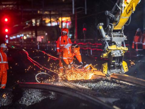 Machine cutting the train track at night. There is sparks coming off the track and 3 workers in personal protective equipment