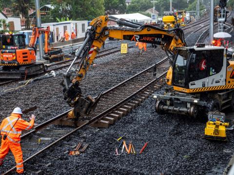 A heavy machine is picking up and moving the rail track. Construction worker in high viz is directing the operator of the machine. 