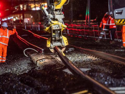The arm of a heavy machine is picking up and moving the rail track at night time. Construction worker in high viz is directing the operator of the machine. 
