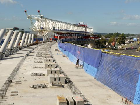An on the ground view looking across at the construction of the railway bridge deck surface with rows of concrete slabs lined up to the right, pillars of concrete to the left and the large white gantry crane in the background.