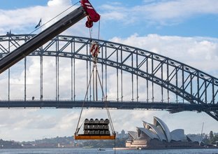 A crane is lowering sleepers for delivery to Blues Point constriction site. The Opera house and Harbour Bridge are in the background.