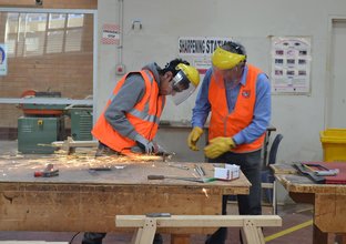 A teacher supports a student while he uses a grinder on metal. Sparks fly in the image as both people are wearing full PPE.