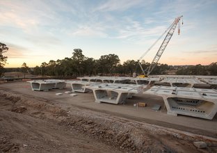 Precast skytrain segments are pictured in a dirt holding yard with a crane in the distance.