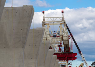 An on the ground view looking up at the gantry crane being lifted into place on top of raised concrete pillars for the construction of Sydney Metro's skytrain. 