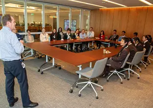 A man is presenting to a several people sitting around an O-shaped table in a board room 