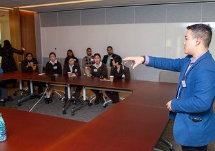 A man is presenting while pointing to several people sitting around an O-shaped table in a board room 