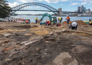 A wide angle of archaeologists in high vis and hard hats working in the excavation area at the Blues Point construction site. Sydney Harbour Bridge and city skyline is seen in the background.