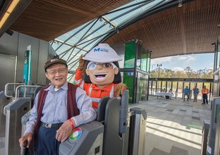 An older gentleman poses with Sydney Metro mascot of a female construction worker outside of the opal card readers at Tallawong Station.