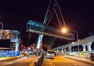 An on the ground view showing a crane lifting a segment of the pedestrian walkway  into place  at night at Sydney Metro's Kellyville Station.