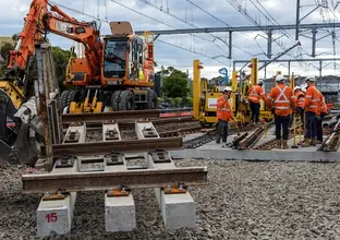 An on the ground view looking at a section of train tracks laid on top of a concrete slab that will be lifted into place by the crane digger with a group of five construction workers in the background working on the track line behind between Sydney Metro's Sydenham and Bankstown Stations. 