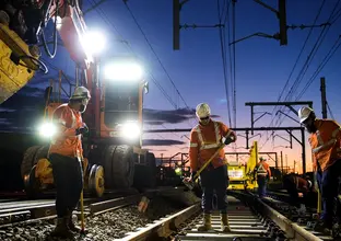 An on the ground view looking at a group of three construction workers laying tracks down on the track line between Sydney Metro's Sydenham and Bankstown Stations  at sundown with the crane digger lights shining bright in the background.