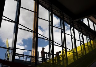 A view from the inside of Sydney Metro Kellyville Station looking up wt two construction workers installing glass windows at the station. 