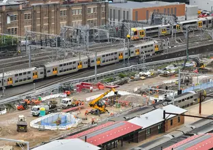 A bird's eye view looking across at the construction site of the Sydney Metro station box at Central Station with many heavy machinery on the ground between the platforms as two trains are leaving the platform in the background.  