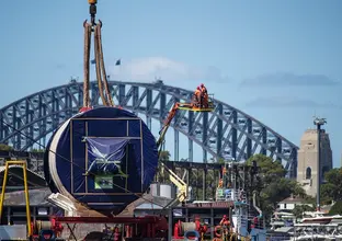 An on the ground view looking across at Tunnel Boring Machine arriving at Sydney Metro's The Bays construction site with shots of the Sydney Harbour Bridge in the background. 