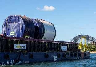 An on the ground view looking across at Tunnel Boring Machine on a boat as it is arriving at Sydney Metro's The Bays construction site with shots of the Sydney Harbour Bridge in the background. 