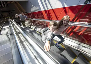 Two children are riding up the escalators smiling with their parents further behind them.