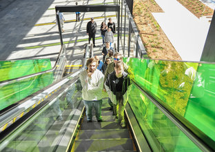 A number of people are riding up green glass-panelled escalators at Rouse Hill Station.