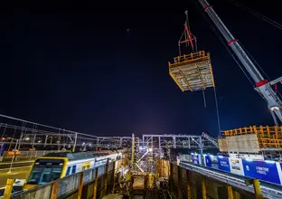 Frame work is lifted into place by a crane on the new concourse as it is being constructed at Sydney Metro's Sydenham Station. The is a train parked in the platform adjacent to the construction site at night time. 