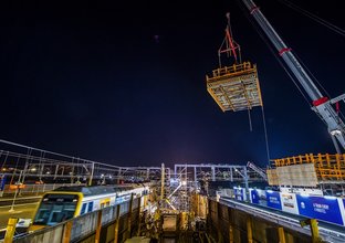 Frame work is lifted into place by a crane on the new concourse as it is being constructed at Sydney Metro's Sydenham Station. The is a train parked in the platform adjacent to the construction site at night time. 