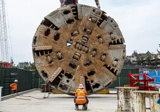 The cutterhead of tunnel boring machine Mum Shirl being lifted from the ground into the air by crane, whilst a construction worker supervises.