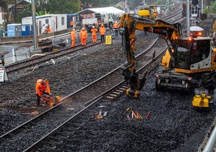 Machine moving the track whilst a construction worker cuts the track. There are several construction workers and shed in the background. 