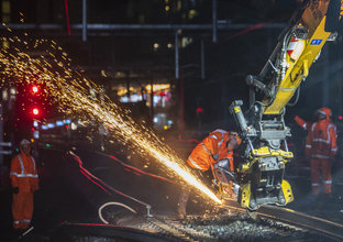 Machine cutting the train track at night. There is sparks coming off the track and 3 workers in personal protective equipment
