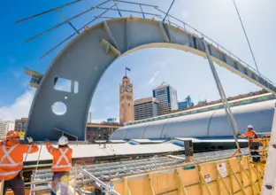 Three construction workers in high viz are working near a large metal lift shaped like a hockey stick. Central Station tower is in the background.