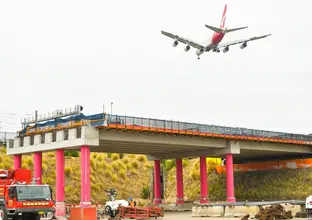 An aqueduct in Sydenham is lifted with a girder lift and sits on pink poles during construction while a plane flies overhead.