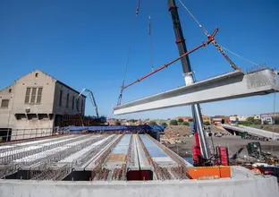 A crane lifting a beam into place at the Sydenham Station and Junction site. There is a building and blue sky in the background.