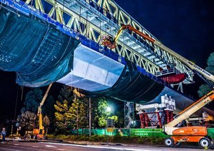 The skytrain over Memorial Avenue is under construction at night with a cherry picker lifting a worker onto the skytrain bridge.