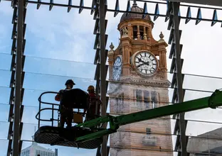 Two workers in high viz are standing on a cherry picker machine, with glass panes of a window behind them. The Central Station clock tower can be seen in the background.