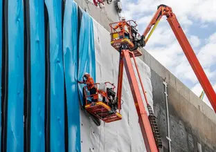 Workers in high-vis are on two cherry picker lifts installing blue waterproof sheeting to the walls of Waterloo Station box.