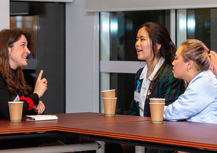 A woman and two students are sitting and talking in a boardroom with takeaway coffee cups sitting on the table.