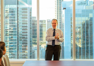 A man in a tie is presenting to people in a boardroom with a city skyline in the background.
