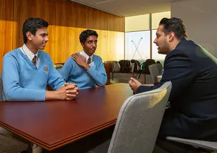 A man and two students in their school uniforms are sitting down in discussion at a boardroom table.