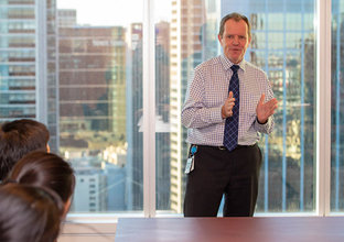 A man in a tie is presenting to people in a boardroom with a city skyline in the background.