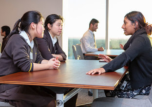 A woman is talking to two students in their school uniforms while sitting down at a table.