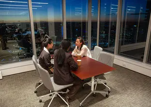 A woman sitting down and talking to two students in their school uniforms at table in a boardroom with a night time city skyline in the background.