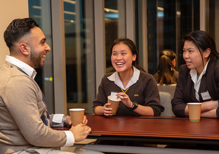 A man is sitting at a table talking and laughing with two students in their school uniforms.