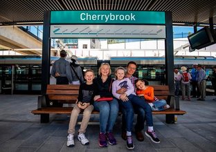 A mother and father pose with their three children sitting on a bench at Cherrybrook station platform.