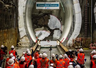 An on the ground shot behind a large group of construction workers watching as Tunnel Boring Machine breaks through the final section of the wall at Sydney Metro's Crows Nest Station. 