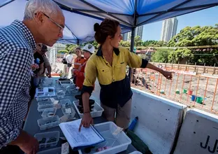 Sydney Metro employees showing community members the archaeological artefacts found at Waterloo Station pointing at a list of artefacts and where on the construction site behind them they were found. 