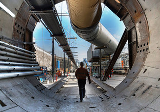 An on the ground view of a construction worker walking through Tunnel Boring Machine (TBM) 2 Florence with a large pipe overhead and construction site in the background. 