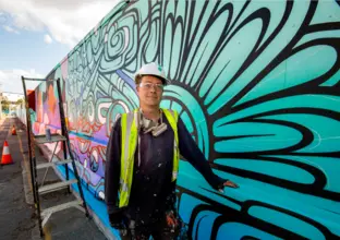 An artist in a high-vis vest and hard hat poses with the mural he has painted at Marrickville Metro Station. A ladder with a spray can on top sits behind him.