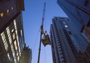 An on the ground view looking up at an excavator is being lifted into place by a crane in between the Sydney sky scrapers in the CBD. 