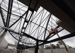 An on the ground view looking up at the steel beam reinforcement for the platform canopy at Sydney Metro's Cudgegong Road Station with a construction worker on a cherry picker carrying out work above. 