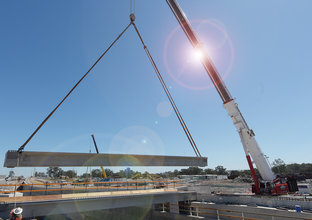 An on the ground view looking cross the construction of the bridge works as a crane is lifting a large piece of concrete material whilst a construction worker guides it into place with a rope at Sydney Metro's Kellyville Station. 