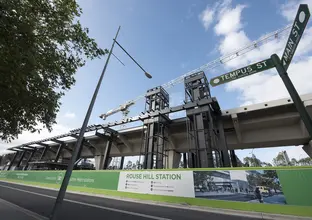 An on the ground view looking up at the station construction bridge with a large white crane in the background at Sydney Metro's new Rouse Hill Station from the corner of Tempus Street and Main Street.