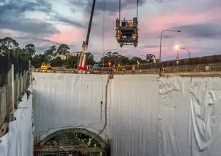 An on the ground view above a tunnel construction site with a crane in the background lifting a concrete paving machine down to the site with construction workers looking on the side guiding the machine operator into place. 