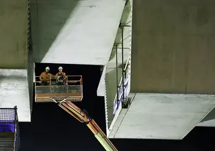 An on the ground view looking up at two construction workers on a crane lift as the final segment is nearing completion. 